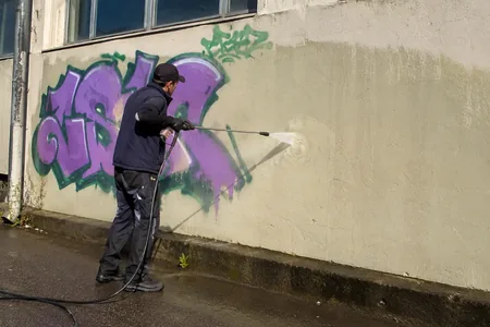 Grafiti eemaldamine survepesuriga, meister peseb lilla ja rohelise grafitiga heledat seina, lahusti kantakse grafitile alt üles, et nõrguv vedelik ei jätaks kuivale seinale triipe.