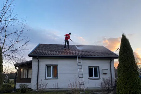 Katuse survepesu, meister seisab heleda puitmaja tumedal katusel survepesuri lantsiga, seinale toetatud redeli ülaots kinnitatakse rihmaga, mis on igas kaubikus kaasas.
